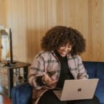 A woman with a curly afro, dressed in a plaid jacket, smiles warmly at her laptop while sitting on a blue couch, illustrating the convenience and support of virtual therapy sessions for Miami Mental Health.