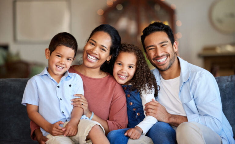 "A joyful family portrait featuring a smiling mother and father with their two young children sitting closely together on a comfortable sofa in a warmly lit living room. The family exudes happiness and warmth, highlighting the importance of mental health and well-being. This image represents the kind of supportive environment fostered by experts like Lazaro Gafas, a dedicated Psychiatric Mental Health Nurse Practitioner who specializes in helping families achieve mental wellness and happiness."