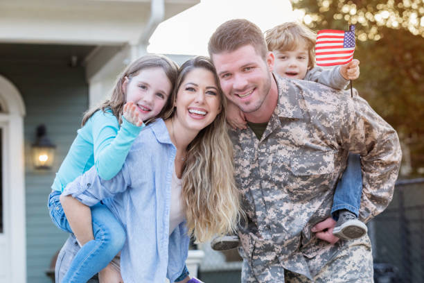 A happy family standing together in front of their home, featuring a father in military uniform, a mother, a young daughter, and a young son holding an American flag. This image represents the importance of mental health services for military families.