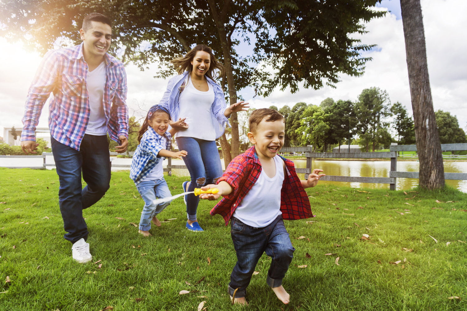 Happy family of four, including two children, running and playing joyfully on a lush green lawn near a serene lake under a sunny sky, promoting the values of Miami Mental Health through vibrant outdoor activity and family bonding