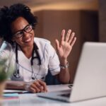 "A cheerful female doctor with curly hair and glasses, wearing a white coat and stethoscope, waves at a laptop screen, representing the accessibility of telehealth services in Miami Mental Health."