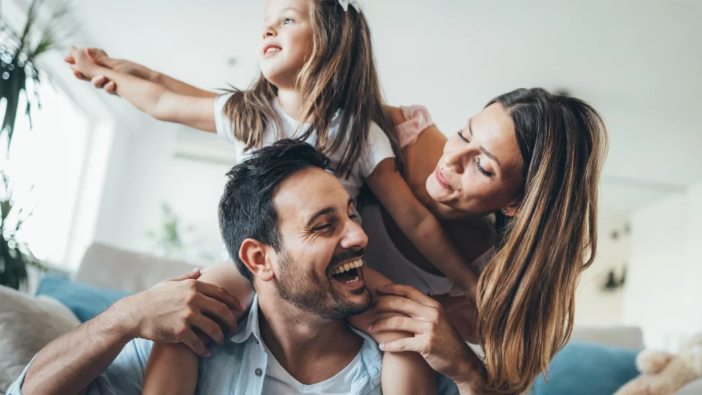 Smiling family at home with father holding daughter on shoulders and mother looking on happily, showcasing the joy and connection fostered through services at Miami Mental Health.